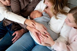 Family hands cradling a sleeping newborn baby together on a couch.