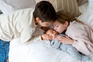 Two children lovingly cuddle with a sleeping newborn baby wrapped in a soft blue blanket on a white bed.