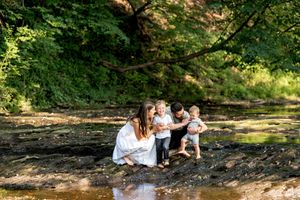 Young family with two toddlers crouching together by a shallow creek in a sunny forest.
