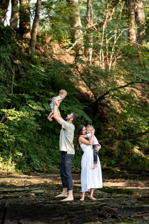 Parents lifting two young children joyfully while standing barefoot on rocks in a green forest.