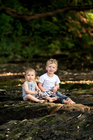 Two young brothers sitting barefoot on mossy rocks in a lush green forest setting.