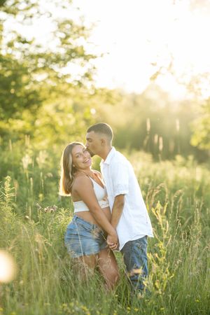Couple sharing a romantic moment in a sunlit meadow during a maternity photoshoot, woman smiling at camera.