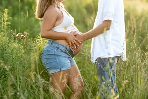 Close-up of expecting couple holding hands over pregnant belly in a sunlit wildflower field.
