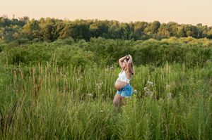 Pregnant woman in denim shorts posing in tall grass during a warm sunset maternity photography session.