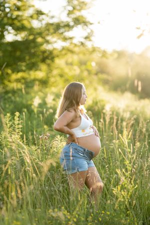 Pregnant woman in denim shorts and floral crop top cradling her baby bump in a sunlit green field.