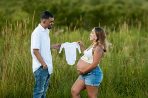 Expecting couple smiling while holding a tiny white baby onesie in a green meadow during maternity shoot.
