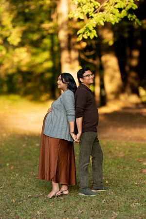 Expecting couple standing back-to-back holding hands on sunlit autumn park path, woman cradling baby bump.