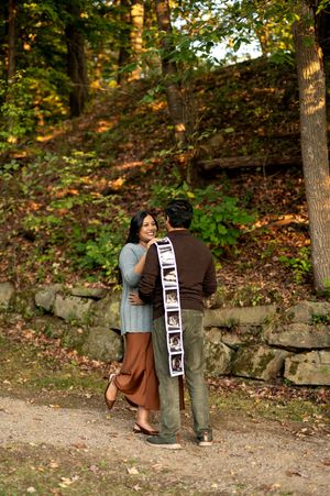 Happy pregnant woman smiling at partner holding ultrasound photo strip in colorful autumn forest.