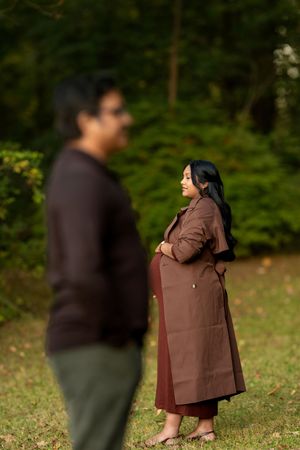 Pregnant woman in brown coat cradling baby bump in lush green park with partner in foreground.