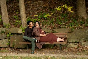 Couple relaxing on wooden bench in autumn forest, pregnant woman in brown dress leaning on partner.