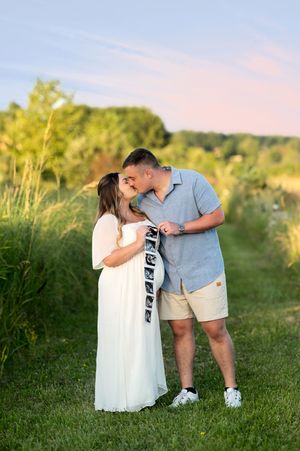 Couple sharing a kiss while holding ultrasound photos during a golden hour maternity shoot in a lush green field.