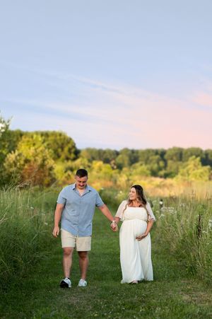 Expecting couple holding hands and walking along a grassy path during a beautiful sunset maternity session.