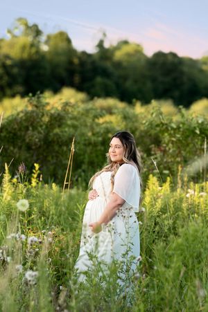 Pregnant woman in white dress cradling baby bump while standing in a wildflower meadow at sunset.