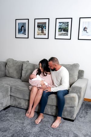 New parents sitting on gray sofa cuddling their newborn baby dressed in pink.