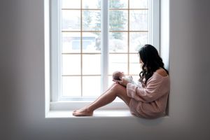 Mother sitting on white window sill cradling her newborn baby in soft natural light.