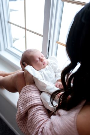 Mother gazing lovingly at her sleeping newborn baby held in her arms by a bright window.