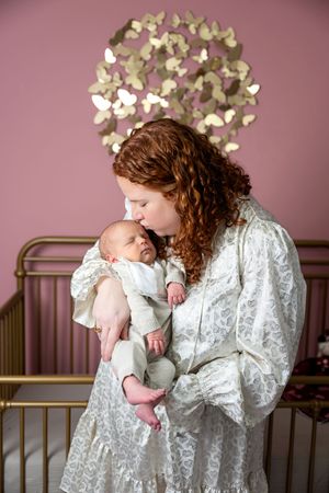 Red-haired mother in a lace gown kissing her sleeping newborn in a pink and gold nursery.