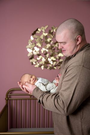 Bald father gently cradling his sleeping newborn baby in a pink nursery with gold butterflies.