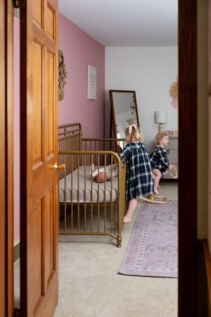 Young girl on rocking horse in a pink nursery room viewed through an open wooden door.