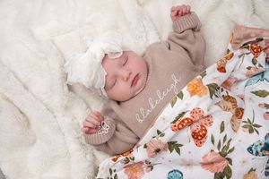 Sleeping newborn baby in a personalized tan gown with a white bow headband on a floral blanket.
