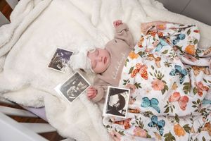 Newborn baby resting beside ultrasound photos on a floral swaddle blanket in a white crib.