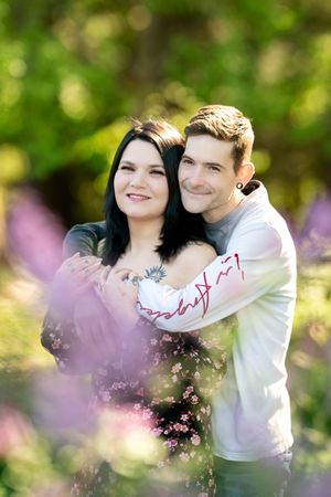 A couple embraces in a sunlit garden surrounded by purple wildflowers, smiling warmly together.