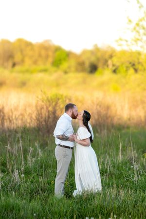 A couple shares a romantic kiss in a golden sunlit meadow, woman wearing a flowing white dress.