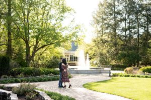 A couple shares a romantic embrace near a garden fountain surrounded by lush green trees.