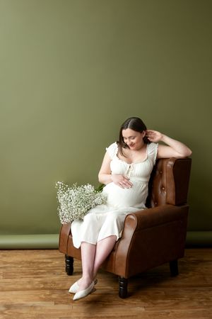 Pregnant woman in white dress sitting in brown leather chair with white flowers.