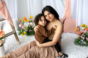 A dark-haired mother and curly-haired toddler share a warm embrace among colorful flowers indoors.