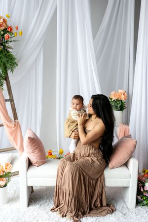A mother in a beige dress holds her baby on a white bench surrounded by flowers and white drapes.