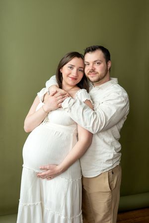 Smiling pregnant couple embracing against olive green wall, woman in white dress.