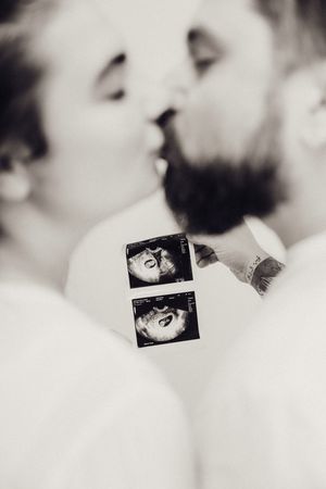 Couple kissing while holding ultrasound images in soft-focus black and white maternity portrait.