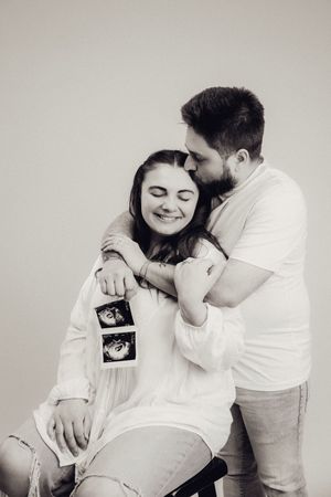 Bearded man kissing smiling pregnant woman holding ultrasound photos in black and white studio shot.