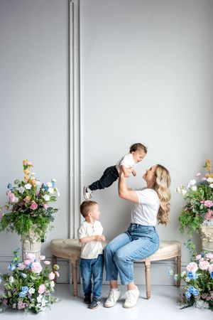 A blonde mother lifts her baby while her toddler son stands beside her on a bench with floral arrangements.