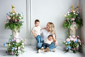 A blonde mother sits on the floor with her two young children surrounded by colorful floral arrangements.