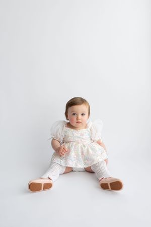 Adorable baby girl in floral dress and white tights sits on white floor looking curiously at camera.