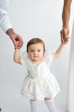 Baby girl in floral dress stands holding parents hands against a white studio background, looking at camera.