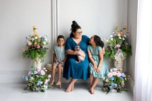 A mother sits with her two children and newborn baby surrounded by colorful floral arrangements indoors.