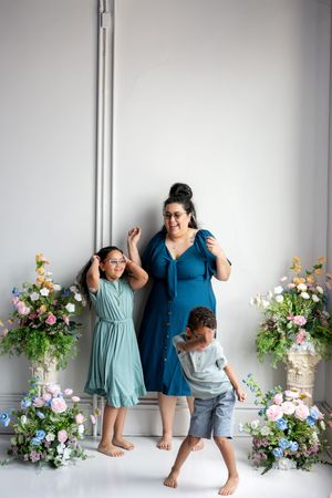 A mother and two children dance and laugh together in front of colorful floral arrangements indoors.