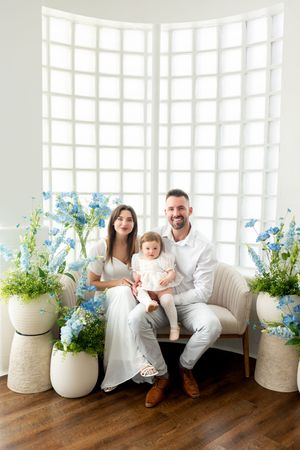 Happy family with baby daughter sits on a loveseat surrounded by blue flowers and white vases in bright studio.