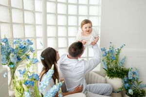 A joyful family photo with parents lifting their smiling baby girl among blue flowers in a bright white room.
