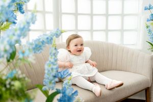 Adorable baby girl in white dress sitting on a beige sofa surrounded by blue flowers in a bright room.