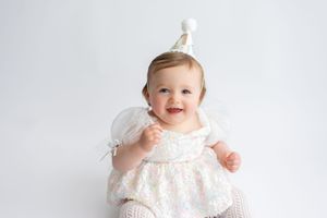 Cute baby girl in white lace dress wearing a party hat smiling against a white background.