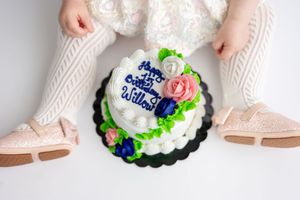 Close-up of a decorated first birthday cake with pink roses and blue flowers between baby's feet.