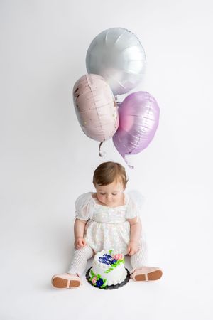 Baby girl in white dress sitting with a birthday cake and pastel balloons on a white background.