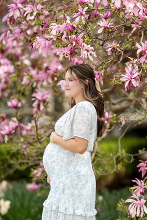 A pregnant woman in a white floral dress cradles her belly beneath a blooming pink magnolia tree.