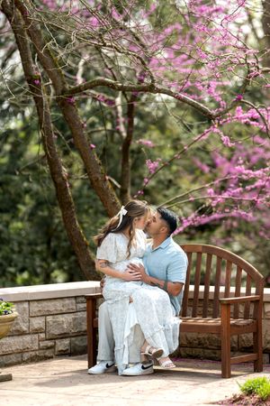 A pregnant couple shares a kiss on a wooden bench beneath a vibrant pink flowering tree.