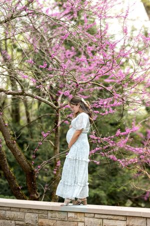 A pregnant woman in a white dress gazes down at her belly beneath a blooming pink redbud tree.
