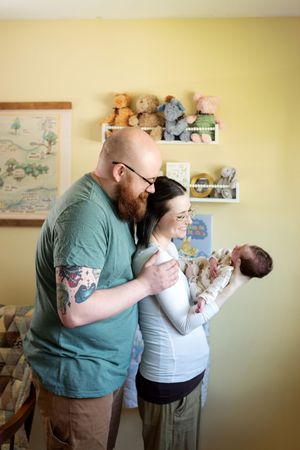 A tattooed father embraces mother holding their newborn baby in a cozy nursery with stuffed animals on shelves.